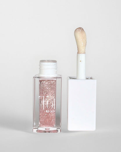 A clear tube with a white cap containing a pink lip oil, alongside a applicator wand with a sponge tip for dry lips. Color Angel